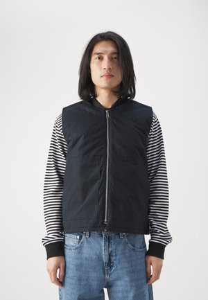 Black vest with a zip front, quilted texture, and two large front pockets, layered over a black and white striped long-sleeve shirt.