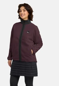 Bordeaux fleece jack met hoge kraag, voorrits en zwarte accenten; gedragen met een gestructureerde zwarte puffer rok en leggings.
