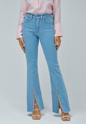 Los jeans de campana de un azul claro tienen un tiro alto, slit en los laterales y bordes deshilachados. Se combinan con sandalias beige con tiras y una blusa rosa.