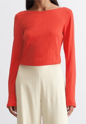 Femme portant un haut rouge moulant à manches longues et un pantalon large crème taille haute sur un fond uni.