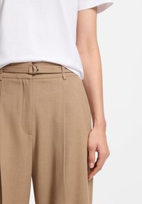 Παντελόνια σε ταμπά χρώμα με λείο ύφασμα, διακοσμημένα με θηλιά και ζώνη. Συνδυασμένα με απλό λευκό T-shirt.