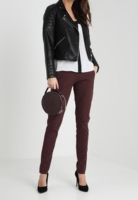 Černá kožená motorkářská bunda, bílá blůza s předním panelem, burgundské slim-fit kalhoty, kulatá vínová kabelka s texturovaným povrchem a černé lodičky.