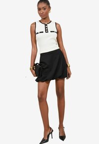 Jupe évasée noire, haut sans manches à rayures blanches avec des accents noirs et des détails de boutons. Modèle tenant un petit sac à main noir texturé.