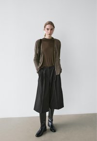 Cardigan marron porté sur un haut marron, associé à un pantalon large noir et des bottes noires élégantes. Textures lisses et tons neutres.