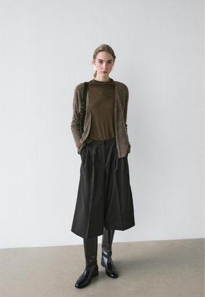 Brauner Cardigan über einem braunen Oberteil, kombiniert mit schwarzen, weit geschnittenen Hosen und eleganten schwarzen Stiefeln. Glatte Texturen und neutrale Farbtöne.