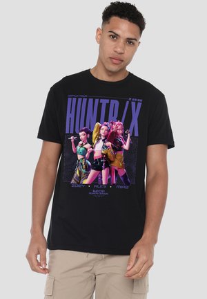 Man wearing black T-shirt featuring three animated female characters in colorful outfits with "HUNTR/X" and tour text on front.
