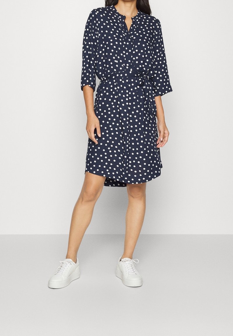 Robe bleu marine à pois blancs, sans col, ceinturée à la taille, manches trois-quarts, associée à des baskets blanches.
