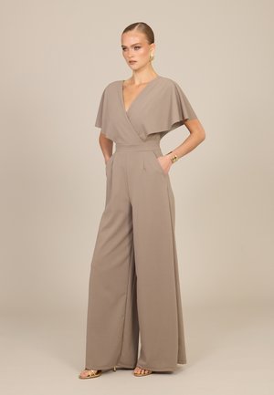 Femme portant une combinaison pantalon taupe à jambes larges avec un profond décolleté en V et de courtes manches cape, debout les mains dans les poches devant un fond uni.