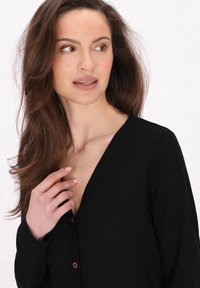 Cardigan nero a maniche lunghe con scollatura a V profonda e bottoni marroni, caratterizzato da una texture morbida che valorizza la figura.