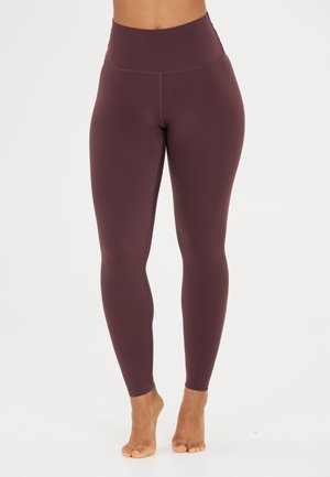 Waist to feet view of person wearing high-waisted, full-length, dark purple leggings standing barefoot on white background.