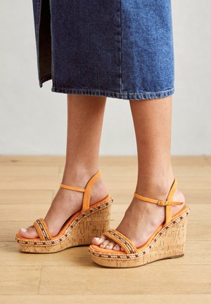 Las sandalias de cuña naranjas cuentan con tiras tejidas y una plataforma con textura de corcho. El diseño incluye una correa en el tobillo con hebilla y puntera abierta.