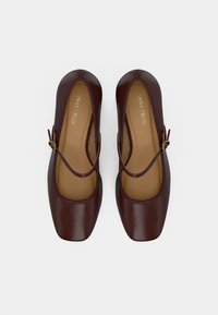 Σκούρα καφέ Mary Jane παπούτσια με τετράγωνα δάχτυλα και ρυθμιζόμενες πλευρικές αγκράφες, προβαλλόμενα από πάνω σε λευκό φόντο.