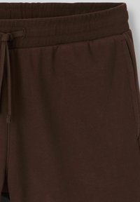 Pantalons de survêtement en coton marron avec une taille côtelée, cordon de serrage réglable et poches latérales. Texture lisse et coupe décontractée.