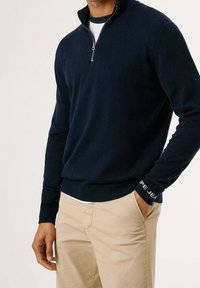 Pull-over bleu marine en mélange de laine avec un col à fermeture éclair quart, des poignets côtelés et un accent de logo sur la manche. Associé à un pantalon beige clair.
