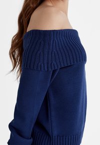Granatowy sweter typu off-the-shoulder z prążkowanym kołnierzem i mankietami, z teksturowanym splotem i luźnym, swobodnym krojem.