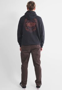 Haut à capuche noir avec un grand design graphique brun au dos, associé à un pantalon marron et des chaussures noires. Les matériaux semblent décontractés et confortables.