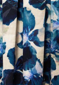 Tessuto floreale con grandi fiori blu e verde scuro su uno sfondo beige chiaro, con una superficie leggermente trama e pieghe drappeggiate.