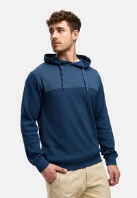 Navyblauwe hoodie met een textuur op het lichaam en een glad paneel in contrast. Bevat een sweatshirtcapuchon met trekkoord en ribgebreide manchetten, weergegeven over lichte broek.