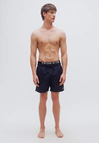 Μαύρο μαγιό σορτς με φαρδιά μέση που φέρει το λευκό κείμενο "CALVIN KLEIN". Το ύφασμα φαίνεται λείο και ελαφρύ. Το μοντέλο είναι χωρίς πουκάμισο.