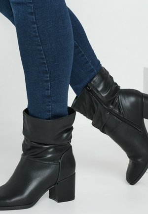 Black faux leather ankle boots with a slouchy design, block heels, and a side zipper, paired with dark blue jeans.