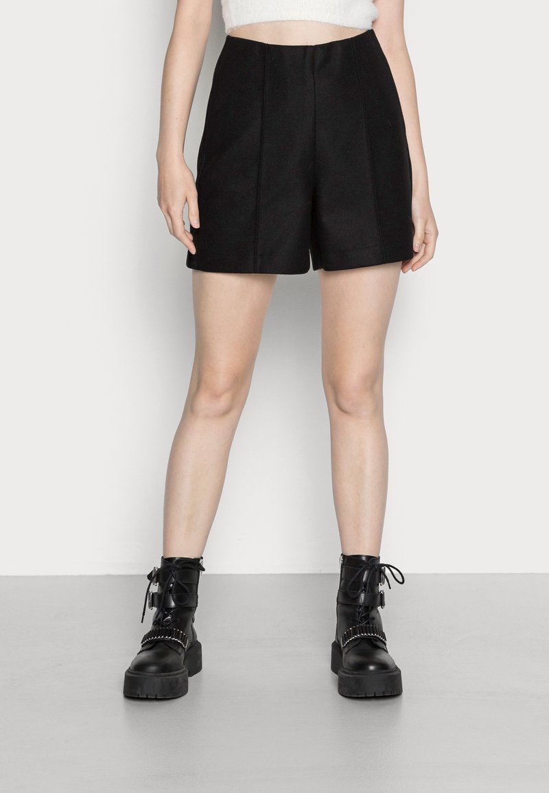 Femme portant un short noir taille haute, un crop top blanc et des bottines noires lacées avec des semelles épaisses, debout sur un sol gris.