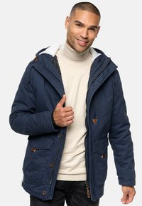 Navyblauwe geïsoleerde jas met een capuchon, voorzien van een faux fur voering, rits- en knoopsluiting, en leren accenten op de zakken.
