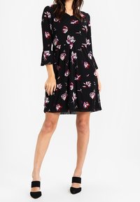 Svart blommig klänning med trekvarts ärmar och trumpetärmar, med en åtsittande överdel, utswingad kjol och vita och lila blommönster.