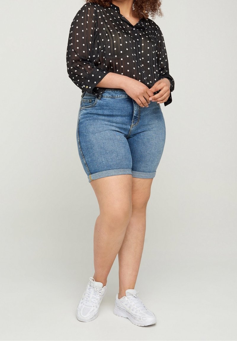 Blusa nera trasparente con pois bianchi, abbinata a pantaloni corti in denim blu con orli arrotolati e sneakers bianche da ginnastica.