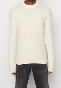 Světlý krémový pletený svetr s kulatým výstřihem, texturovaným vzorem, dlouhými rukávy a žebrovanými manžetami a lemem. Kombinován s tmavě šedými džínami.