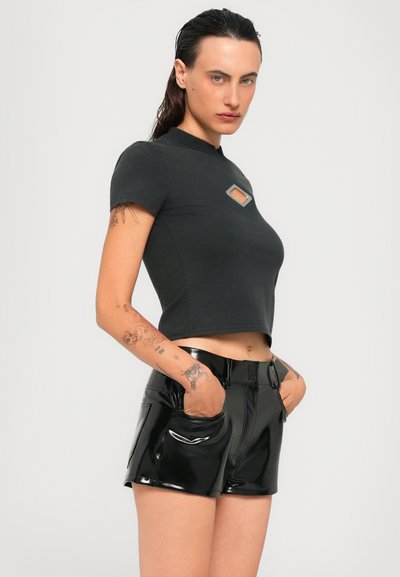 Donna con capelli bagnati che indossa una maglietta nera corta con taglio e pantaloni corti neri alta vita lucidi, in piedi con le mani in tasca su uno sfondo semplice.