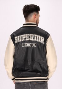 Schwarze und cremefarbene Bomberjacke aus Leder; mit "SUPERIOR LEAGUE" Aufdruck auf dem Rücken; gestreifter, gerippter Kragen und Bündchen.