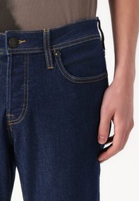 Jeans din denim de culoare albastru închis, cu cusături galbene, închidere cu button și buzunare laterale. Materialul are o textură clasică și netedă.