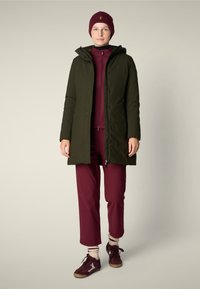Parka verde oliva con capucha, sudadera cremosa con cremallera color burdeos, pantalones burdeos a juego, calcetines de rayas crema y zapatillas marrones con acentos burdeos.