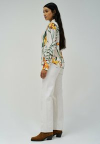 Blusa floreale a maniche lunghe con motivi arancioni e verdi, abbinata a pantaloni bianchi a gamba larga e stivali con tacco marroni. Tessuto morbido.