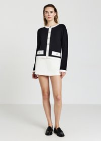 Schwarze und weiße, taillierte Strickjacke mit goldenen Knöpfen, ausgestattet mit zwei vorderen Taschen. Ein weißer Minirock rundet das Outfit ab, kombiniert mit schwarzen Slippern.