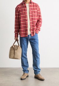 Czerwona i czarna koszula w kratę flanelową, beżowa koszulka, niebieskie dżinsy, jasnobrązowe zamszowe buty, trzymając jasnobrązową torbę z brązowymi akcentami.