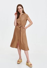 Beżowa, lekko dopasowana sukienka z kołnierzykiem, z zapięciem na guziki z przodu, krótkimi rękawami i wiązanym pasem. Gładki materiał, długość do kolan z rozcięciem po bokach.