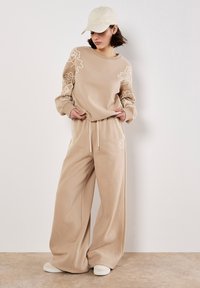 Conjunto de sudadera beige y pantalones de pierna ancha con bordado floral, cintura con cordón y tejido suave. Combinado con una gorra de color claro.