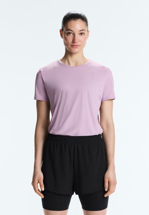 Nő áll előre nézve, világoslila rövid ujjú ingben és fekete réteges rövidnadrágban, egyszerű fehér háttér előtt.