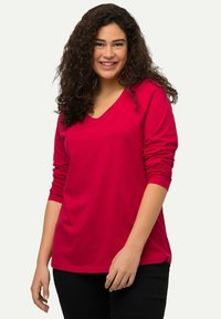 Langärmliges T-Shirt mit V-Ausschnitt aus leuchtend roter Baumwolle, mit lockerem Schnitt und gerippten Bündchen. Kombiniert mit schwarzen Hosen, die eine glatte Textur aufweisen.