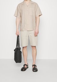 Linnen overhemd met korte mouwen in beige, beige shorts, zwarte geweven sandalen en een zwarte schoudertas met een schouderriem. Eenvoudige, casual outfit.