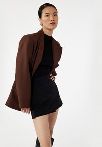 Bruine oversized blazer over een zwarte aansluitende turtleneck en mini-jurk, met zwarte kniehoge laarzen, tegen een effen witte achtergrond.
