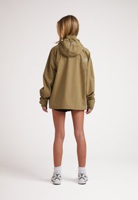 Giacca impermeabile verde oliva con cappuccio, vestibilità rilassata, polsini elasticizzati e dettaglio con logo sulla parte superiore della schiena. Adatta per l'uso all'aperto.