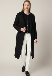 Czarny wełniany płaszcz z dużymi klapami, na pojedynczy guzik, nałożony na czarny kardigan ze złotymi guzikami, w zestawieniu z białymi spodniami i czarnymi butami.