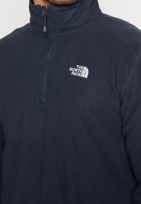 Pullover in pile di colore navy con colletto alto, zip a metà lunghezza e logo ricamato sul lato sinistro del petto. Texture morbida e bordi privi di cuciture.