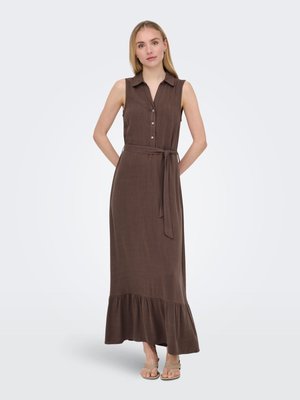 Femme portant une robe longue marron sans manches avec des boutons, une ceinture à la taille et un ourlet volanté, se tenant devant un fond clair uni.