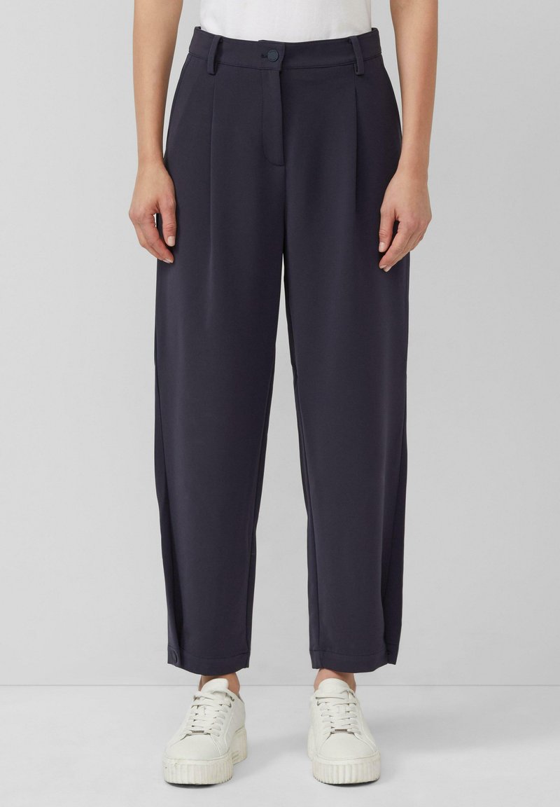 Marineblauwe wijde broek met hoge taille, geplooide voorkant en zijzakken, gecombineerd met witte sneakers voor een casual look.