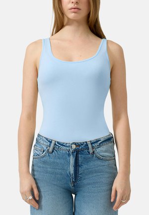 Vrouw draagt een lichtblauw mouwloos hemdje, ingestopt in een hoge taille blauwe spijkerbroek met ringen aan haar vingers.