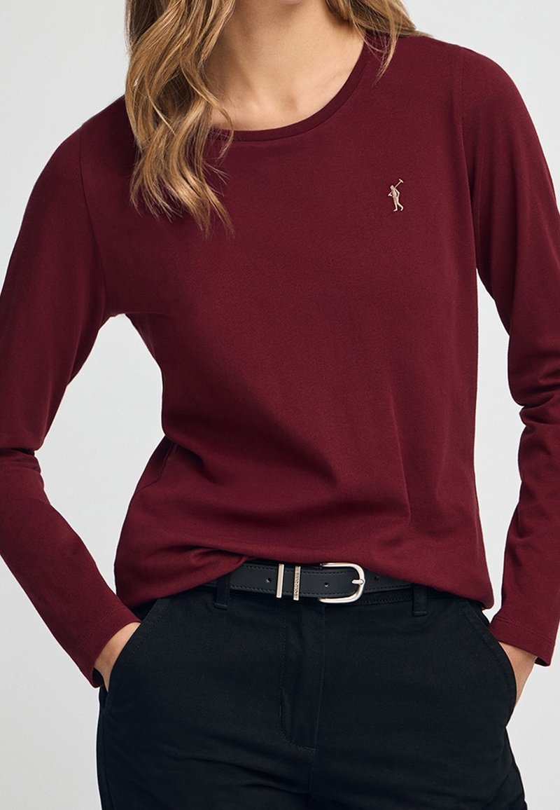 Femme portant une chemise bordeaux à manches longues avec un petit logo joueur de polo sur la poitrine, pantalon noir et ceinture noire, mains dans les poches.