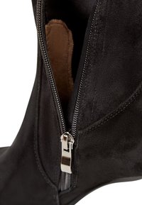 Bota de ante negra hasta el tobillo con cierre lateral de cremallera, que presenta una textura suave y un forro interior marrón contrastante. Detalle de la cremallera en color plata.